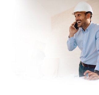 Smiling construction manager in hard hat talking on phone while reviewing blueprints at construction site