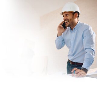 Smiling construction manager in hard hat talking on phone while reviewing blueprints at construction site